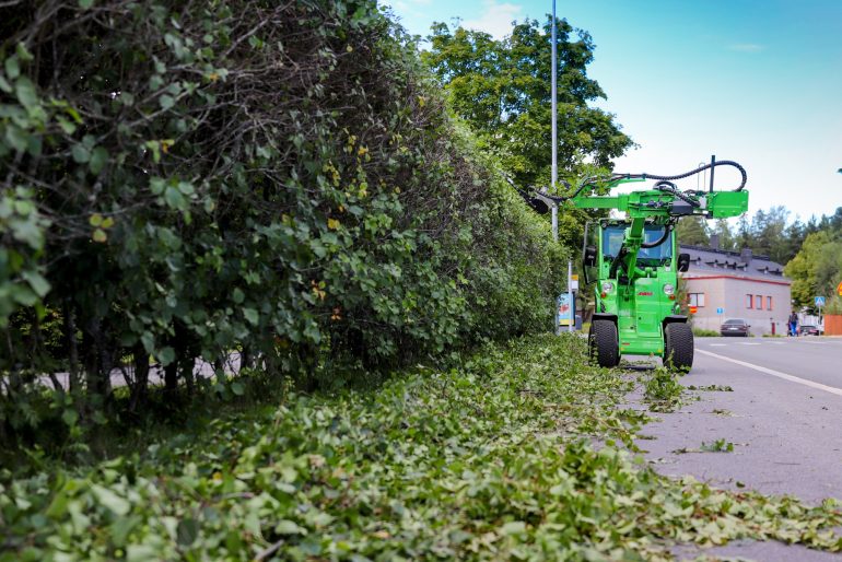 A compact loader cutting a hedge.